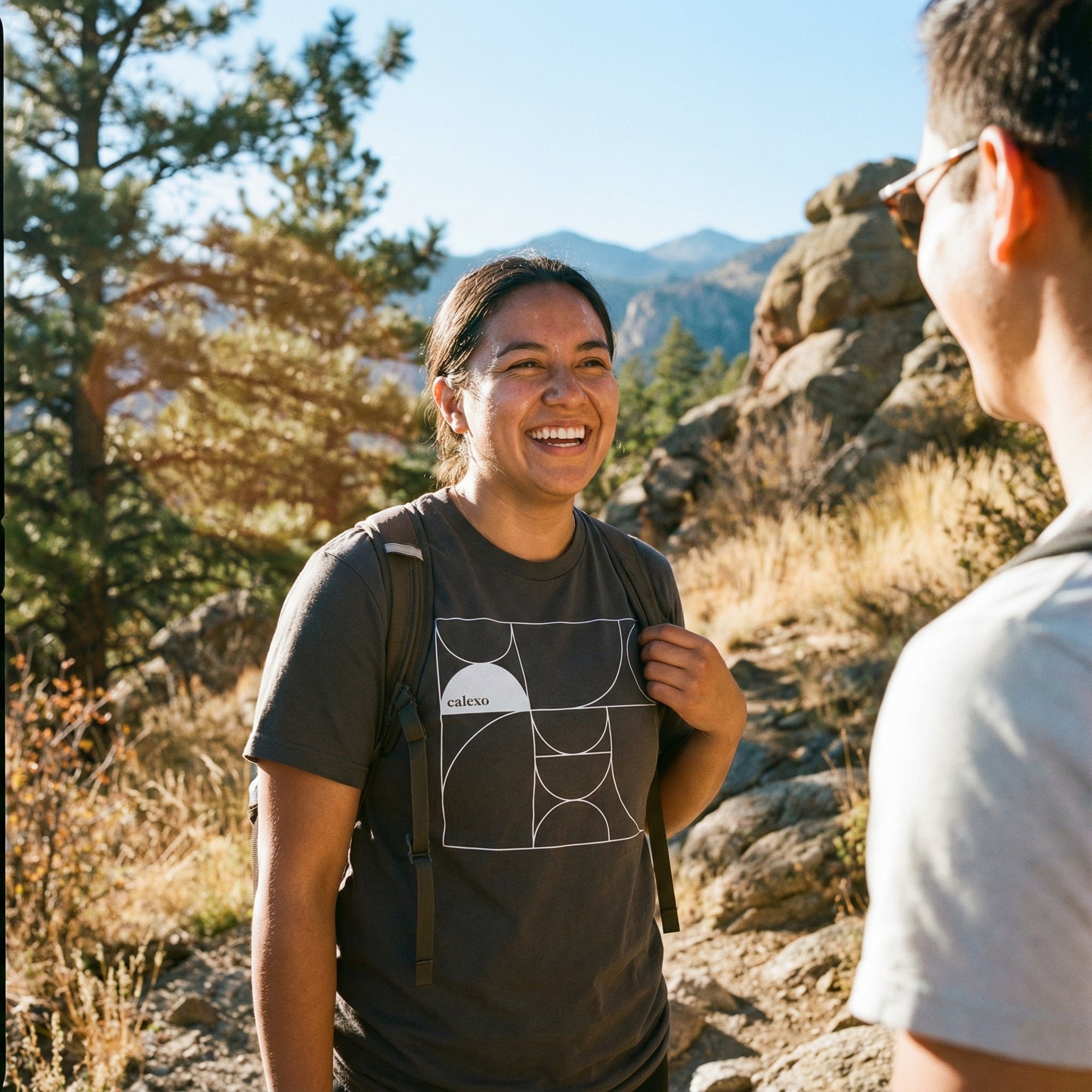 Two people hiking in a mountainous area with trees and rocks in the background.