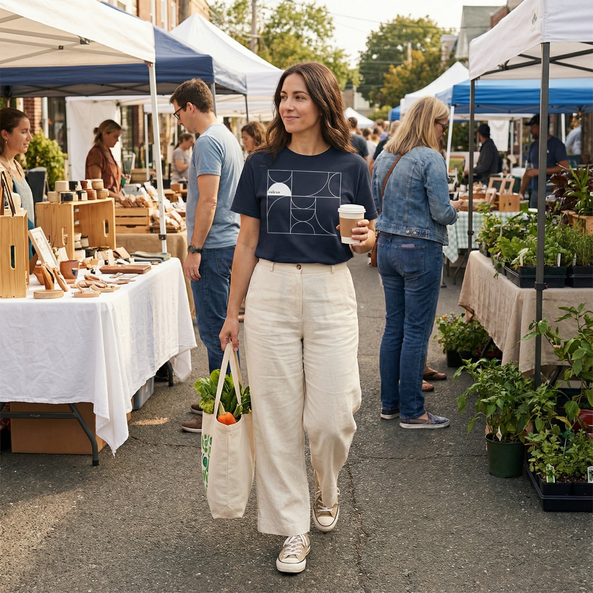 Woman walking at an outdoor market holding a coffee cup and a reusable bag with groceries.