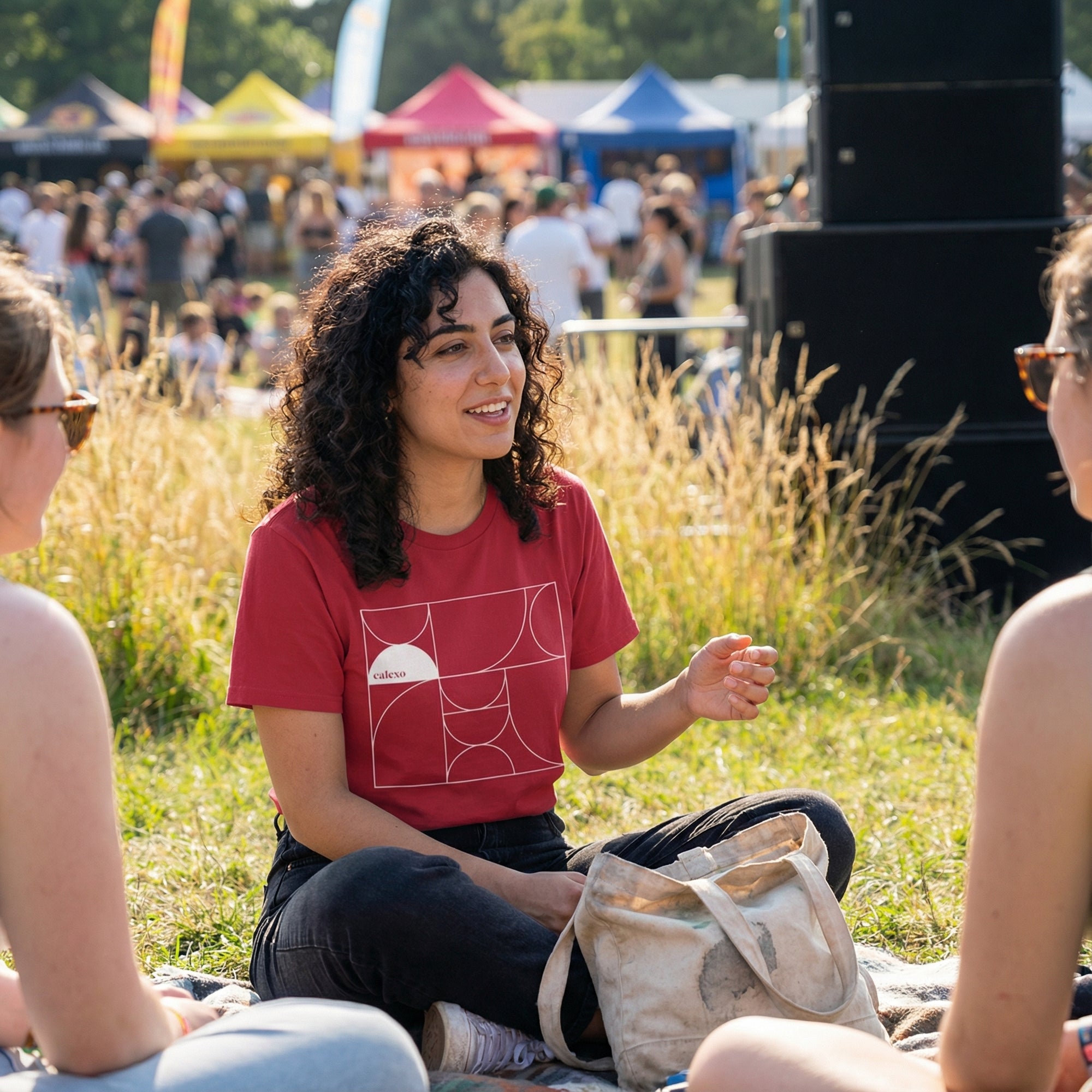 Woman in a red shirt sitting on grass at an outdoor event with a crowd and tents in the background.